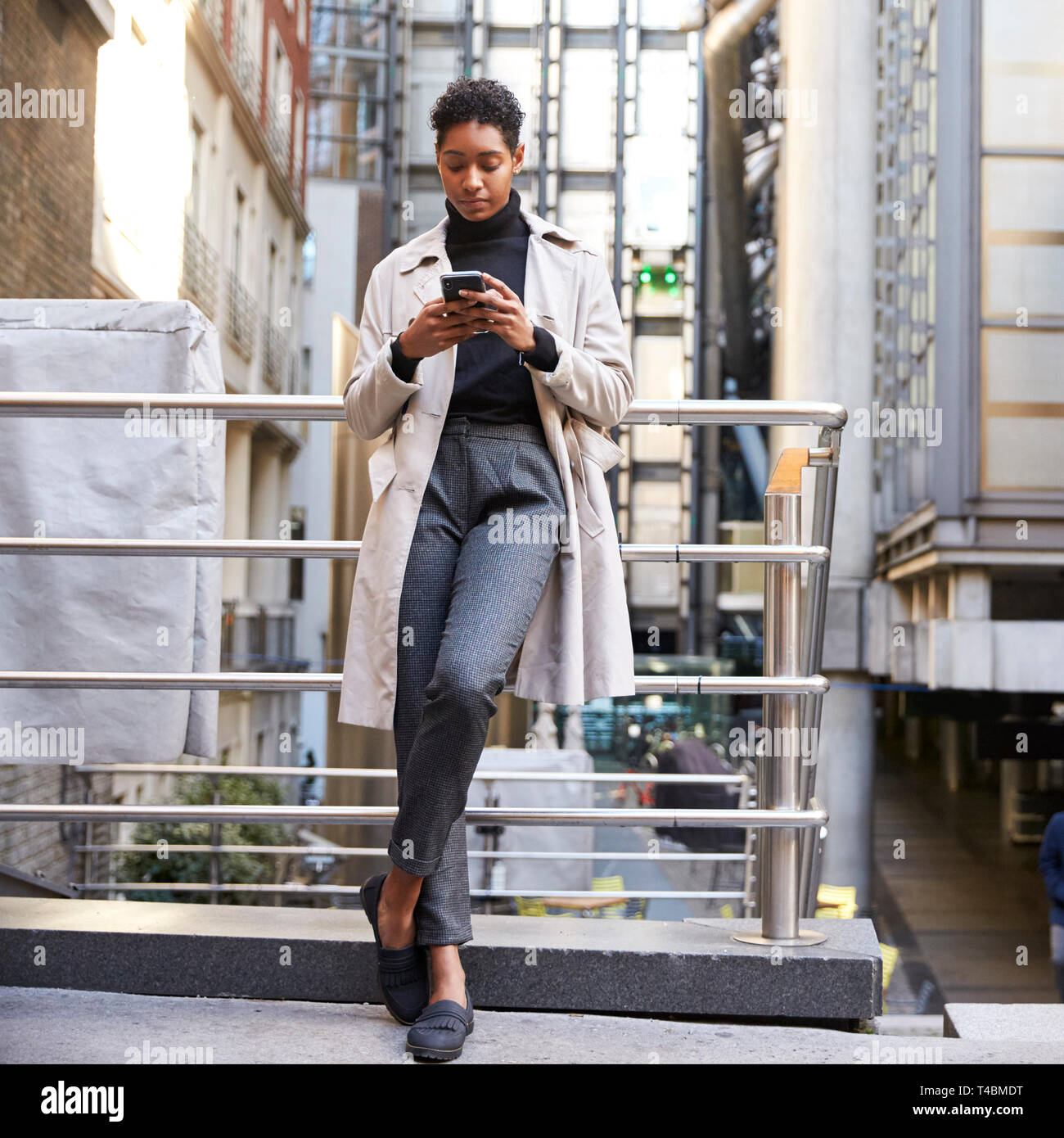 Modische junge schwarze Frau, die in der Stadt lehnte sich auf eine Hand mit Ihrem Smartphone, volle Länge Schiene, niedrigen Winkel Stockfoto