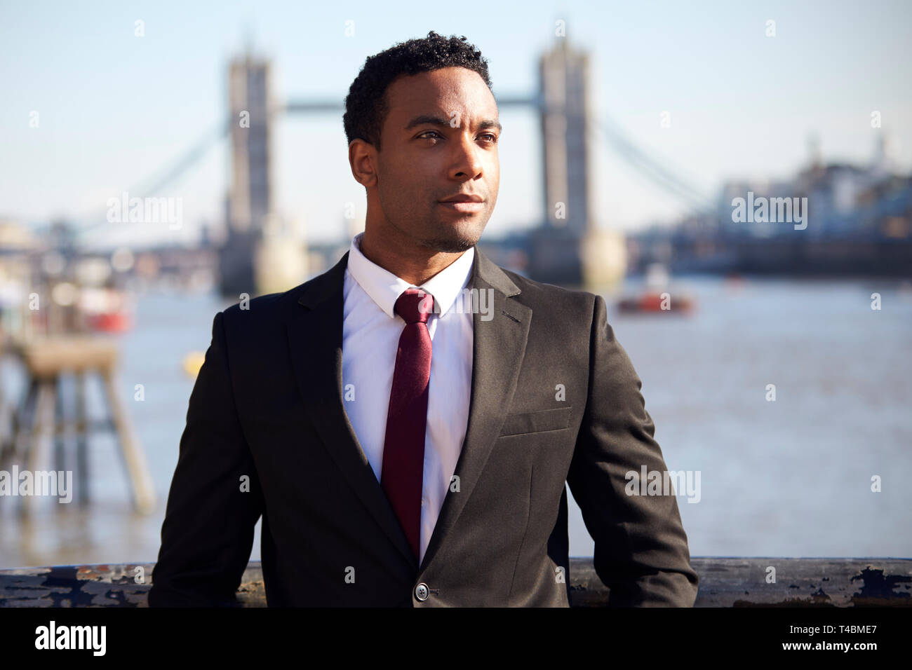 Tausendjährige schwarzer Geschäftsmann mit schwarzen Jacke, weißes Hemd und Krawatte an der Themse Riverside in London, Tower Bridge im Hintergrund, im Vordergrund Schwerpunkt Stockfoto