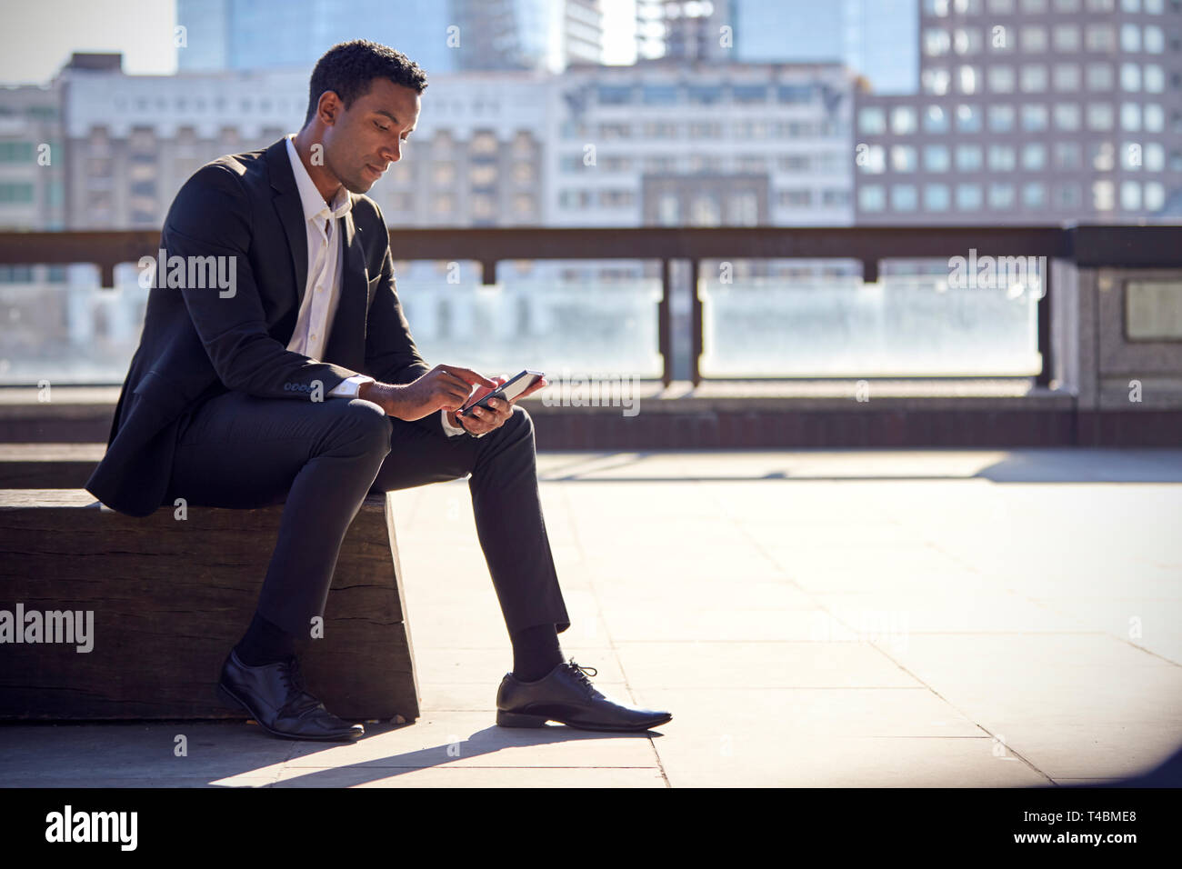 Tausendjährige schwarzer Geschäftsmann mit schwarzen Anzug und weißes Hemd am Ufer der Themse, London sitzend, mit Smartphone, mit Hintergrundbeleuchtung Stockfoto