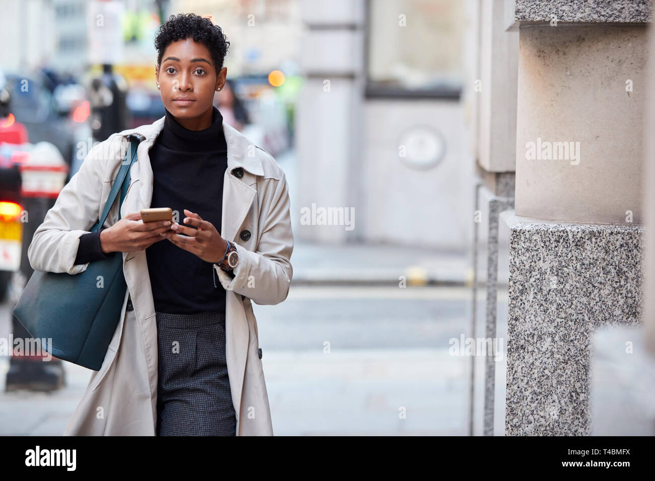 Junge schwarze Geschäftsfrau zu Fuß auf der Straße in London können Sie über ihr Smartphone, Vorderansicht Stockfoto