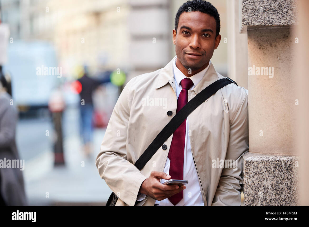 Junge schwarze Geschäftsmann lehnte sich an eine Wand in der Straße in London holding Telefon und lächelnd in die Kamera, selektiver Fokus Stockfoto