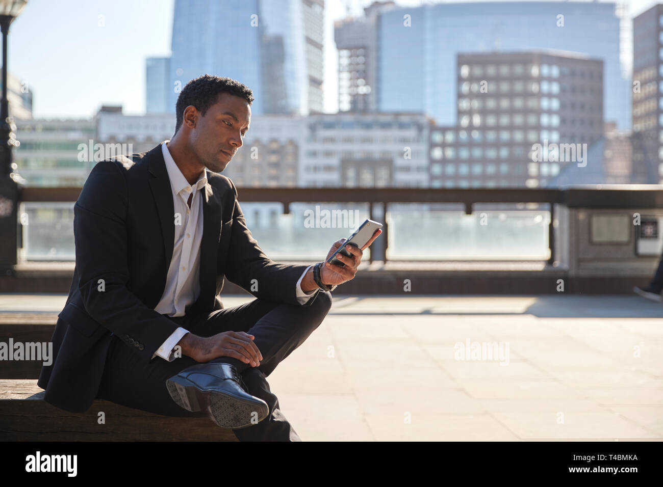 Tausendjährige Geschäftsmann mit schwarzen Anzug und weißes Hemd sitzen auf der Themse Ufer mit Smartphone, in der Nähe Stockfoto