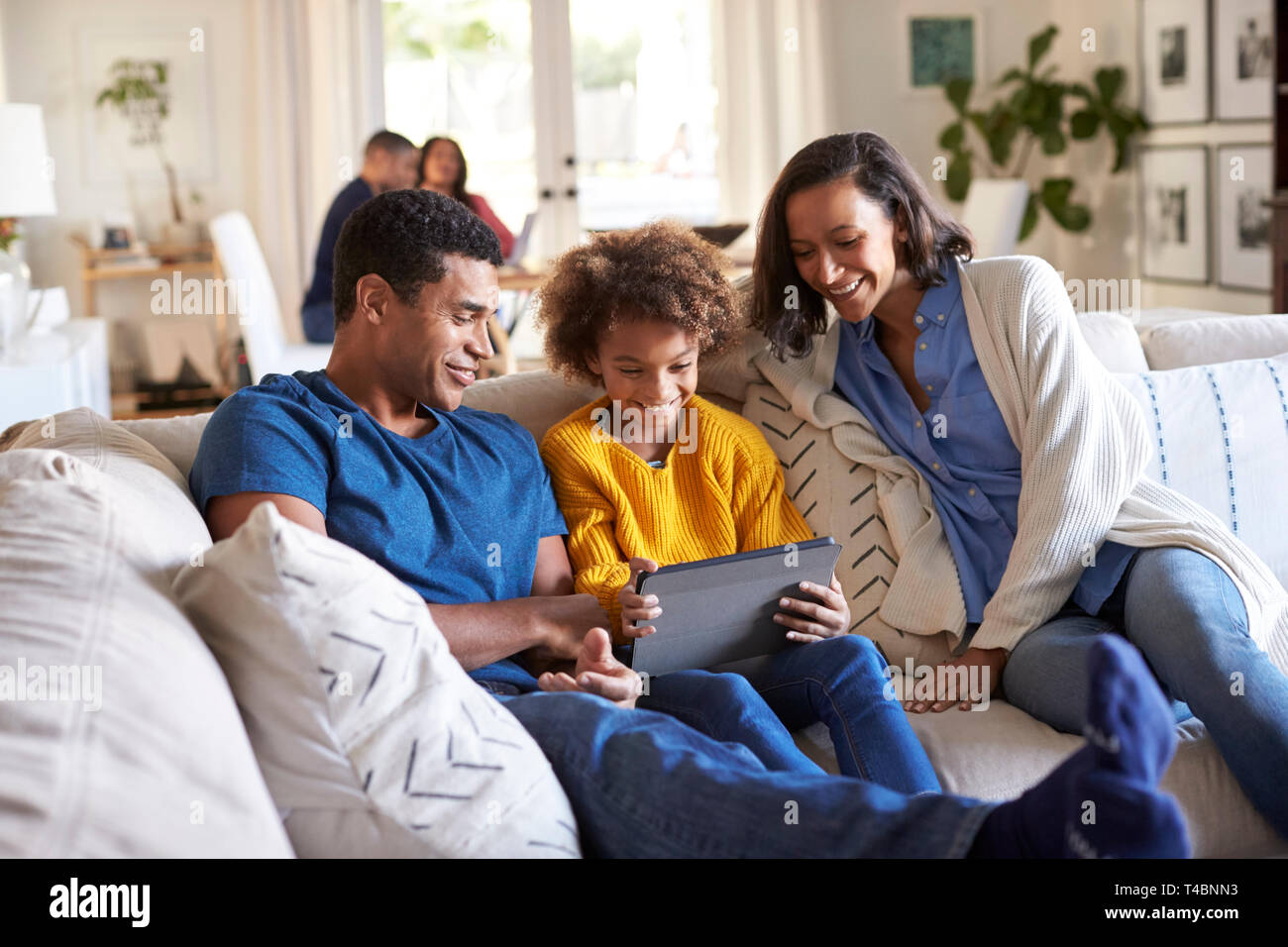 Junge Eltern und ihre vor - jugendlich Tochter sitzt auf einem Sofa im Wohnzimmer mit Tablet-PC zusammen, Großeltern an einem Tisch im Hintergrund sitzen Stockfoto