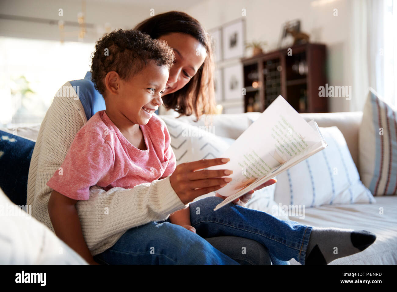 Nahaufnahme der jungen Mutter sitzt auf einem Sofa im Wohnzimmer ein Buch lesen mit Ihrem Kleinkind Sohn, der sitzt auf ihrem Knie, Seitenansicht Stockfoto