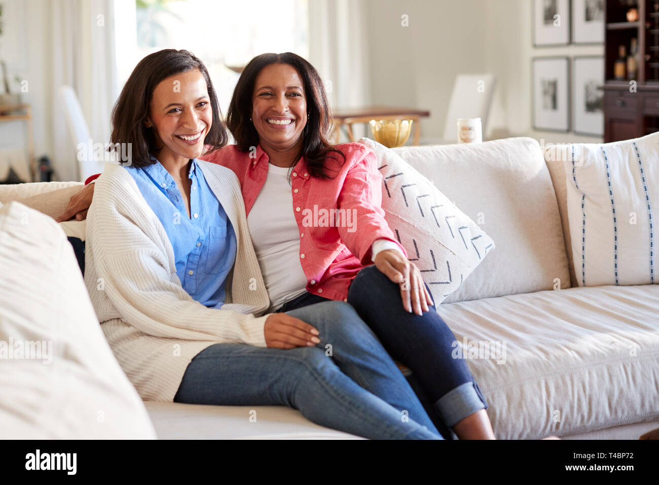 Junge Frau sitzt auf dem Sofa im Wohnzimmer mit ihrer Mutter lächelnd in die Kamera, drei Viertel Länge Stockfoto