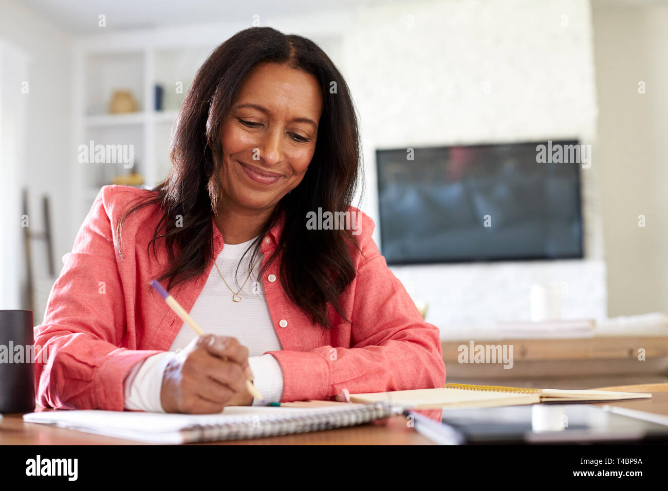 Lächelnd gemischten Rennen pensionierte Frau an einem Tisch schreiben in ihrem Esszimmer sitzend, Nahaufnahme, niedrigen Winkel Stockfoto
