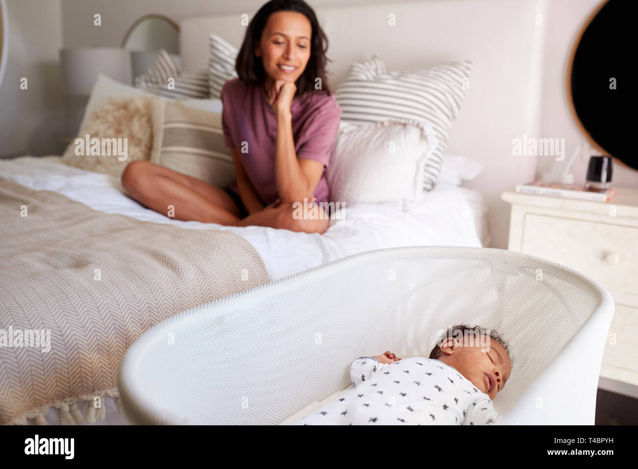 Junge Erwachsene Mutter sitzt im Schneidersitz auf ihrem Bett, ihr drei Monate alten Baby schlafend in seinem Kinderbett, Nahaufnahme Stockfoto
