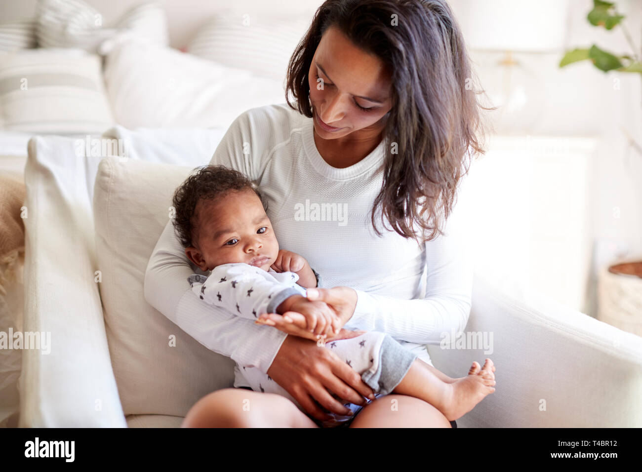 Junge Erwachsene Mutter sitzt in einem Sessel in Ihr Schlafzimmer, Ihr drei Monate alten Baby Sohn in die Arme und schaut ihn an, in der Nähe Stockfoto