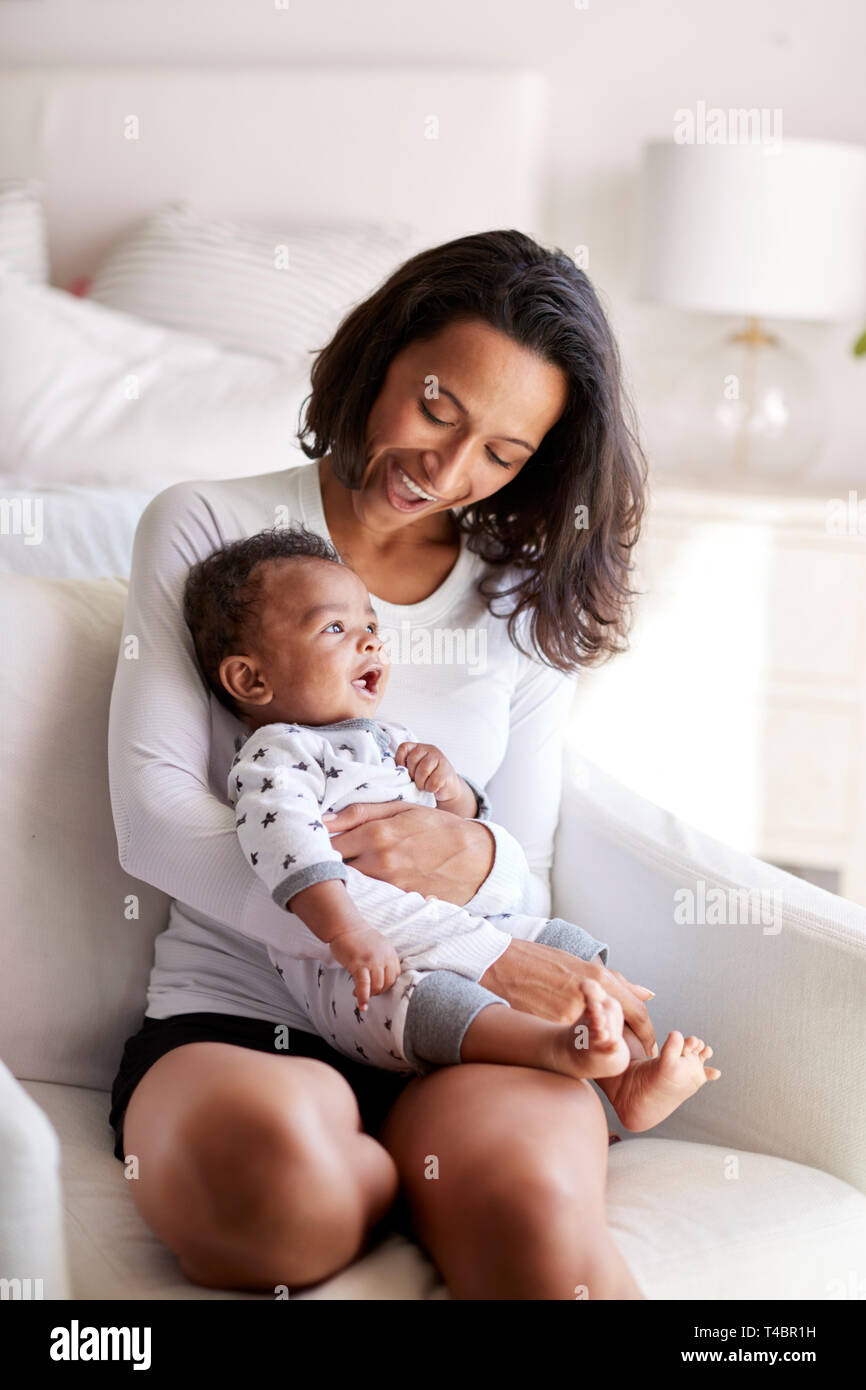 Junge Erwachsene Mutter sitzt in einem Sessel in Ihr Schlafzimmer, Ihr drei Monate alten Baby Sohn in ihre Arme und unten auf ihn schaut lächelnd, vertikal Stockfoto