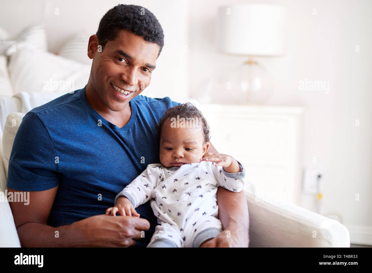 Nahaufnahme von jungen Erwachsenen schwarzen Vaters in einen Sessel seine drei Monate alten Baby Sohn Holding sitzt und lächelt in die Kamera, Vorderansicht Stockfoto