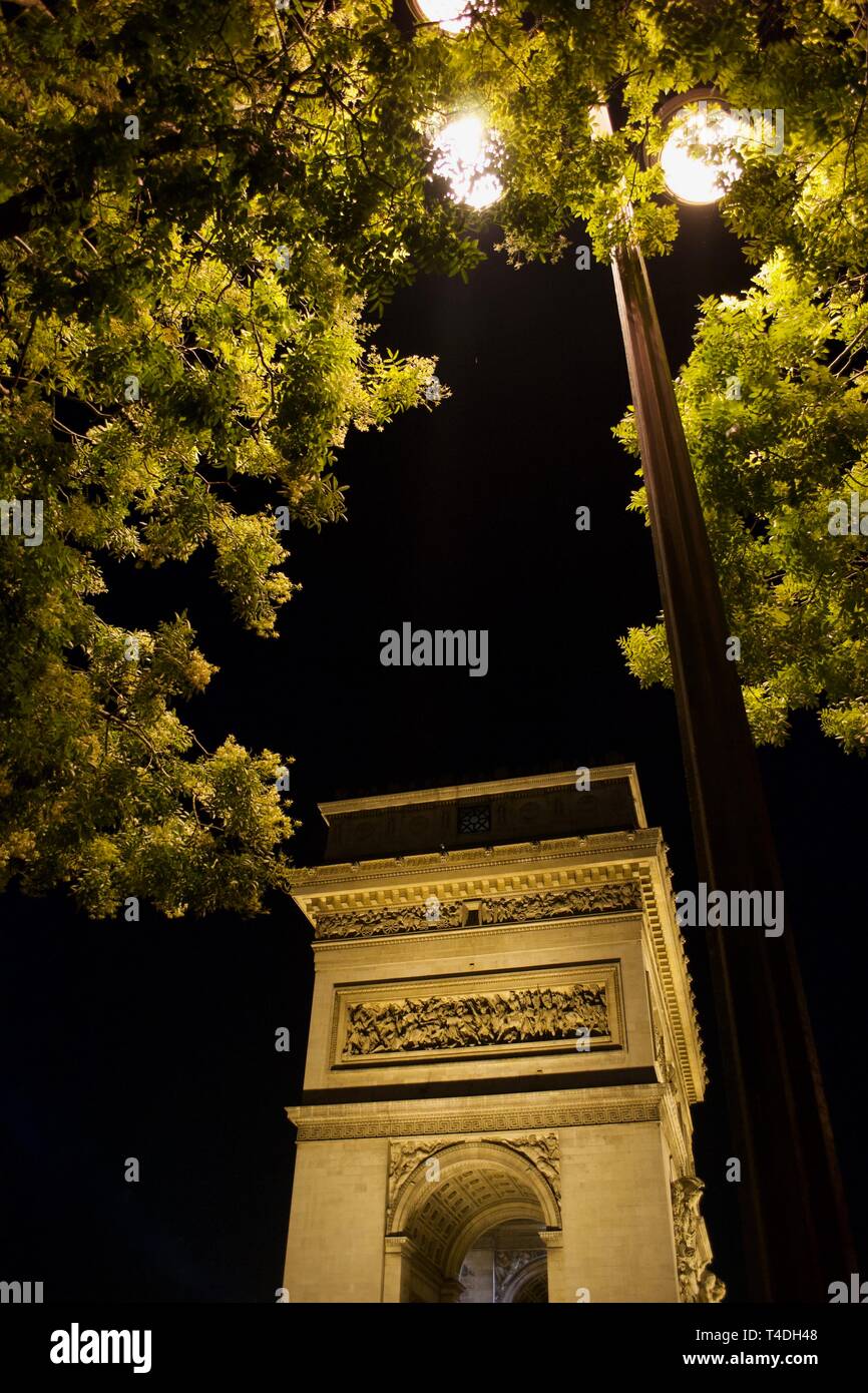 Lampost leuchtet in der Nacht, eingehüllt in Grün der Bäume. Arc de Triomphe leuchtet in gelbes Licht, durch Blätter eingerahmt. Stockfoto