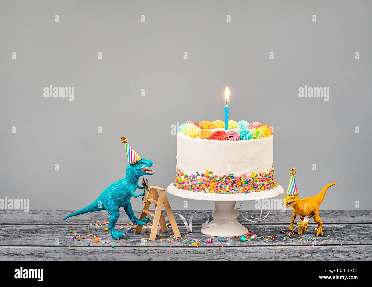 Hungrig Spielzeug Dinosaurier Hüte tragen und halten Gabeln neben einer Geburtstagstorte auf grauem Hintergrund Stockfoto