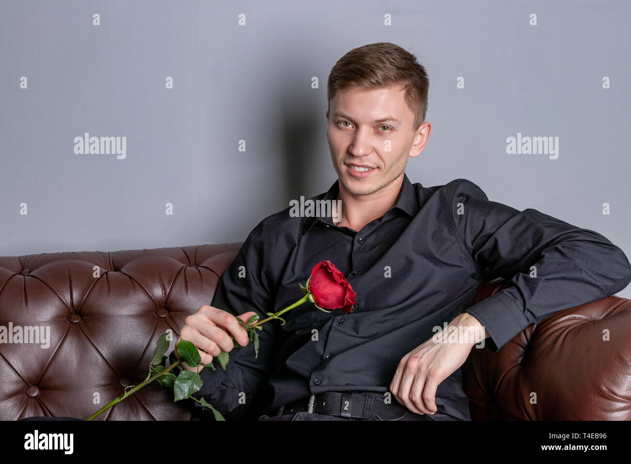 Stattlicher Mann mit schwarzen Hemd sitzt auf dem Ledersofa Holding eine Rose Stockfoto