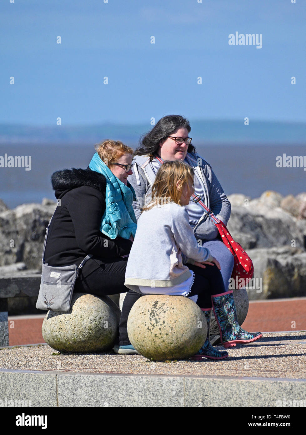 Drei Frauen sitzen auf Stein sitzen, in der Frühlingssonne. Stein, Mole, Promenade, Morecambe, Lancashire, England, Vereinigtes Königreich, Europa. Stockfoto