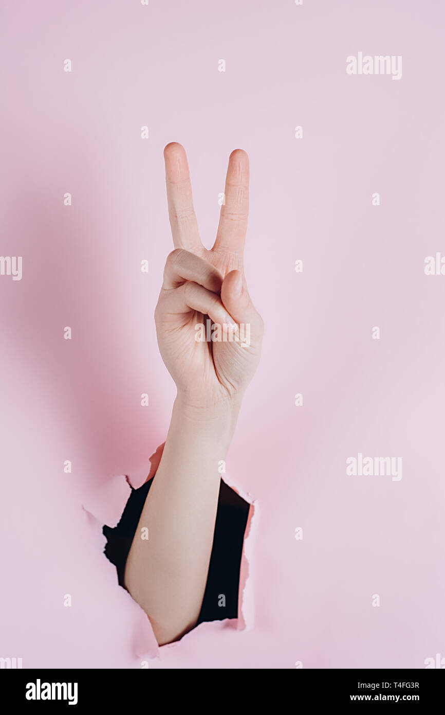 Weibliche zeigen Hände mit Sieg Zeichen, dass durch ein Loch in rosa Papier gesehen werden kann. Körpersprache Konzept. Stockfoto