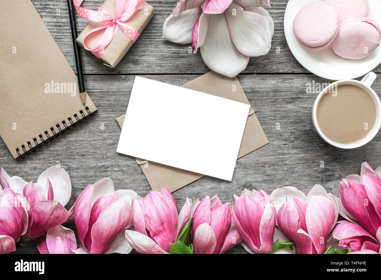 Leere Grußkarte, Tasse Cappuccino, Makronen, Geschenkbox, Notizbuch aus Papier und Magnolia Blumen über rustikalen Holztisch festlichen Hintergrund. Flach Stockfoto