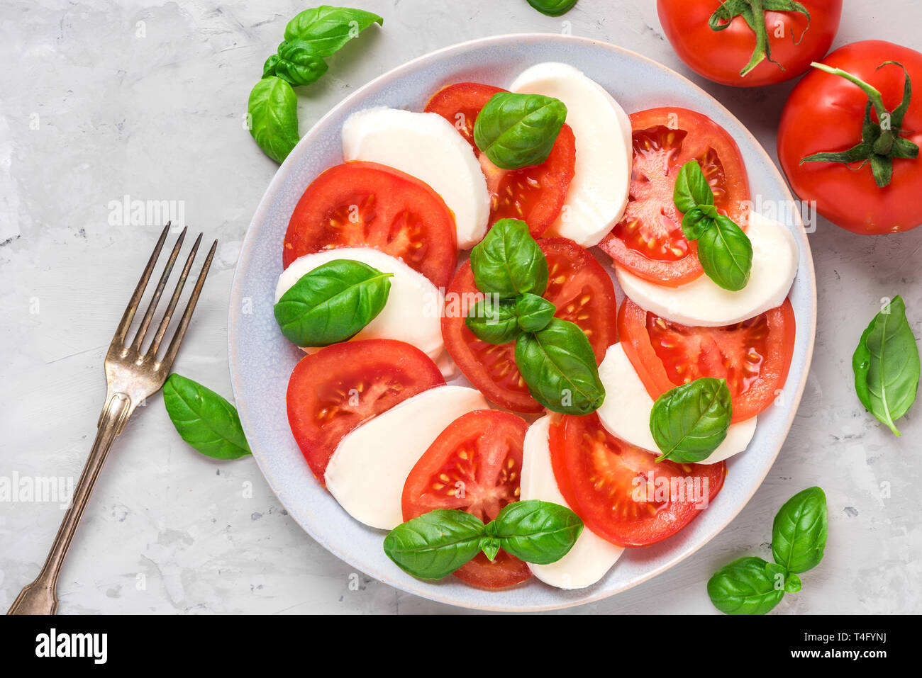 Frische italienische Caprese Salat mit in Scheiben geschnittenen Tomaten, Mozzarella, Basilikum, Olivenöl und Gabel in eine Platte auf grauem Beton Hintergrund. Flach. nach oben v Stockfoto