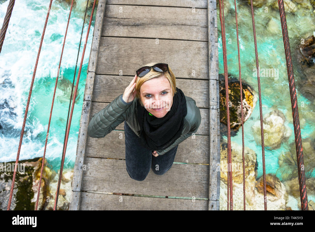 Glücklich lächelnde und entspannende Frau auf einem Tagesausflug auf eine alte hölzerne Hängebrücke über die fließende kristallklare türkisfarbene Fluss Soca, Slowenien Stockfoto