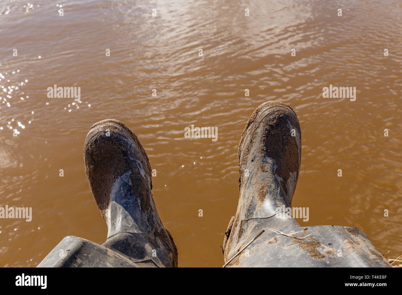 Beine von einem Fischer in schmutzigen Gummistiefel sitzt am Ufer des Flusses mit schmutzigem Wasser im Hintergrund Stockfoto