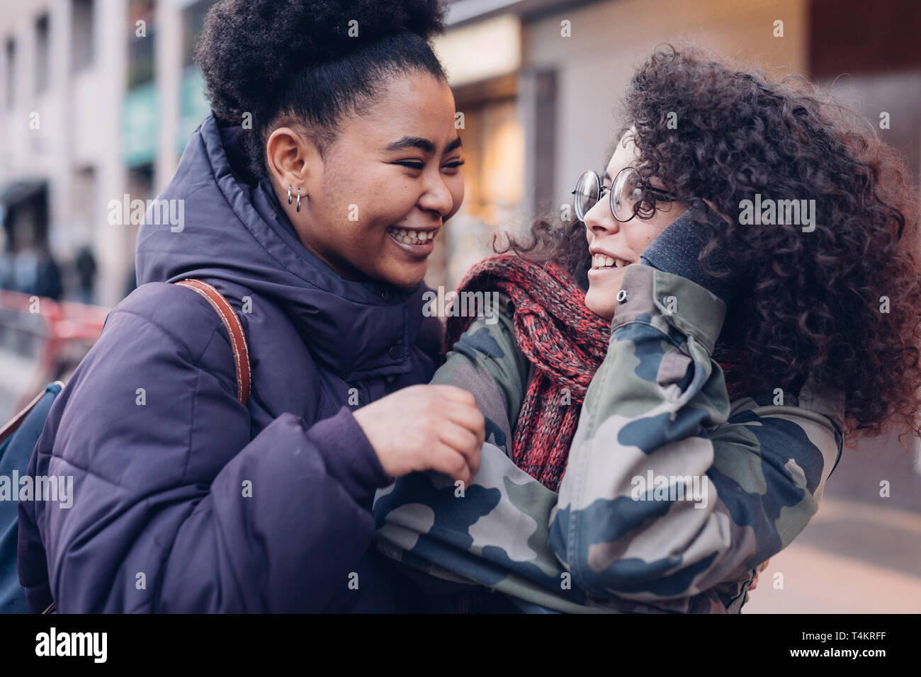 Zwei junge Frauen umarmen in der Straße mit dem lockigen Haar - Freundschaft; heiter; Freude; Stockfoto