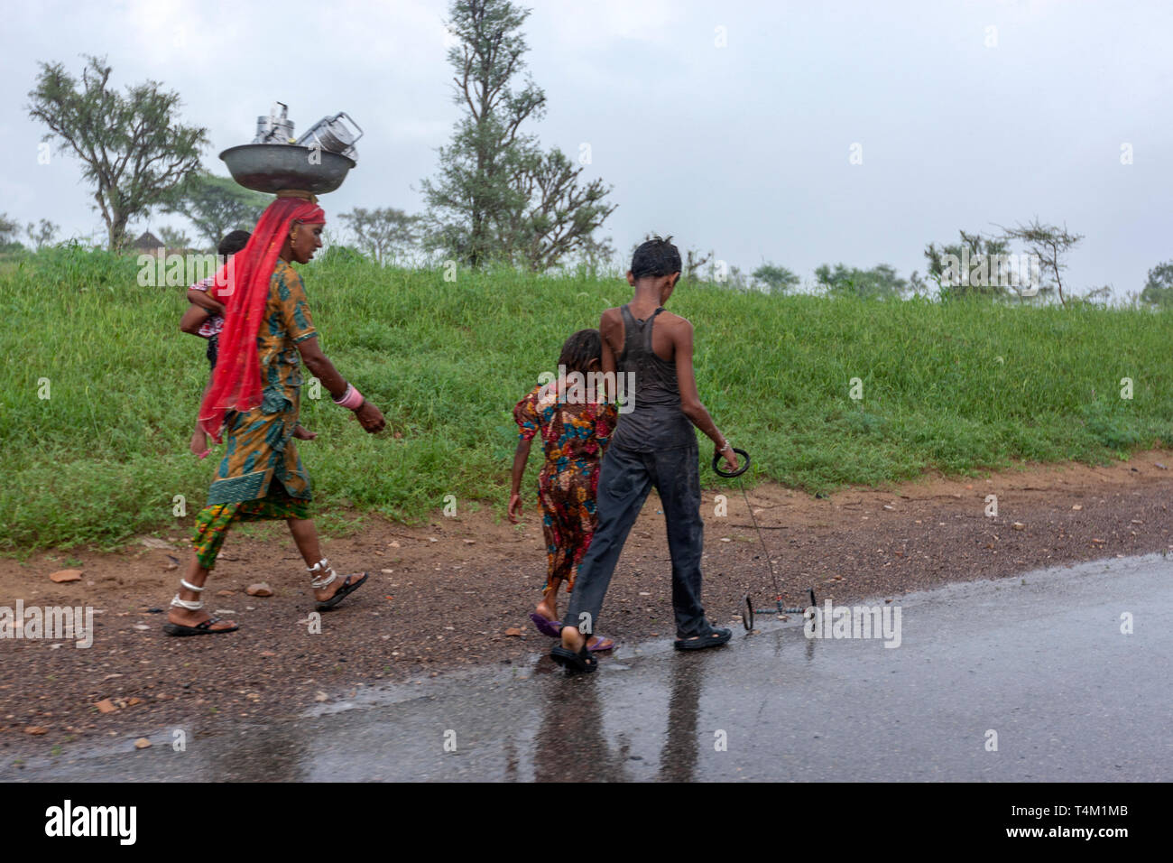 Rajasthani Frau mit seiner Familie zu Fuß entlang einer Straße unter einem starken Regen mit seinem Sohn spielen mit einem rudimentären verdrahtet Auto Spielzeug in Rajasthan, Indien Stockfoto