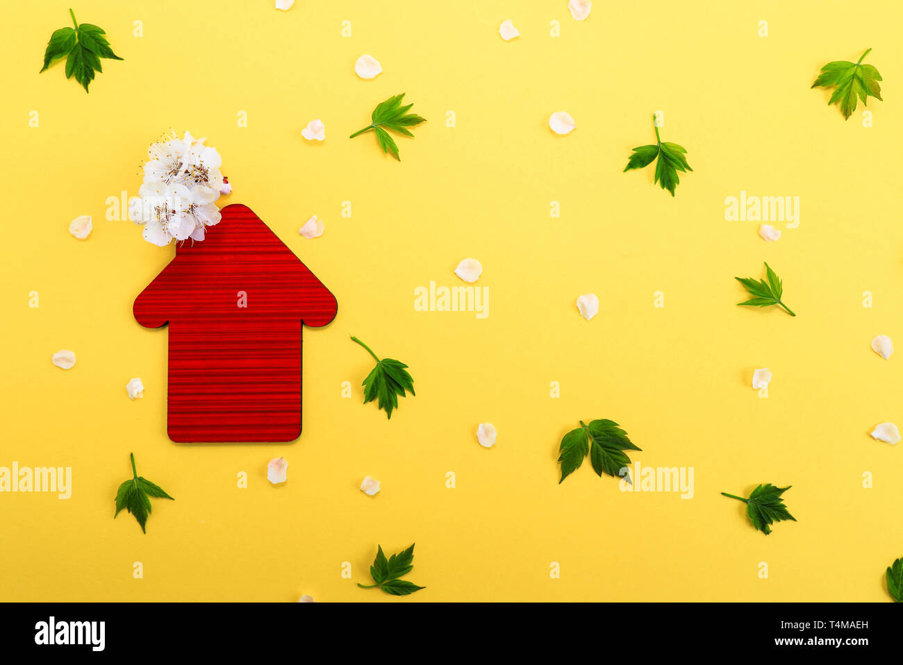 Red toy House, weißen Blüten auf gelbem Hintergrund mit grünen Blättern. Immobilien Geschäftskonzept, Platz für Text, bezahlbaren Wohnraum für Familien Stockfoto
