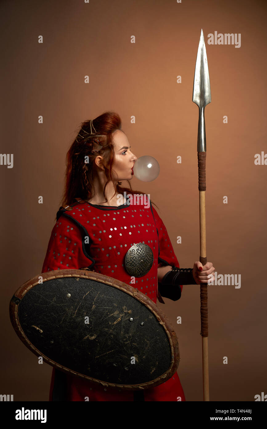 Tapfere weibliche Krieger tragen in Rot mittelalterlichen Kostüm blasen große Blase von Kaugummi. Schön, wunderschöne Frau mit roten Lippen im Studio Posing, Schild und Speer. Stockfoto