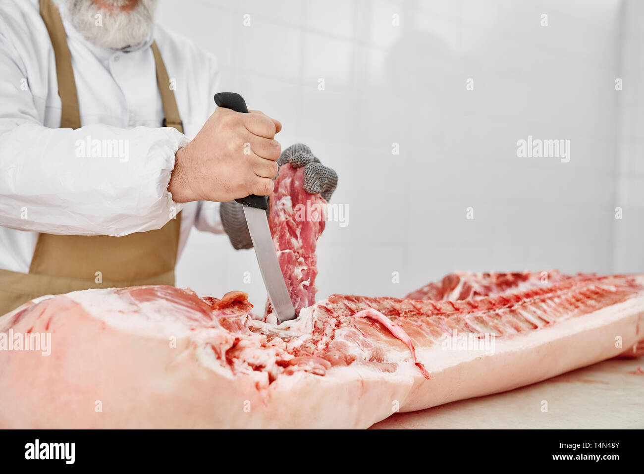 Nahaufnahme von Raw gehacktes Schweinefleisch Schlachtkörper und männlichen Händen mit Messer. Metzger in weißer Uniform und braune Schürze in Fleisch shop arbeiten, Schneiden frisches, rohes Fleisch. Fleisch Fertigung. Stockfoto
