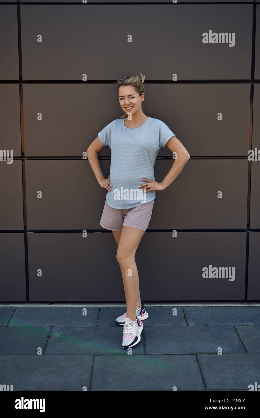 Fit athletische junge schwangere Frau in Sportkleidung vor einem Geschäftshaus mit Händen auf den Hüften in die Kamera lächelnd posiert Stockfoto