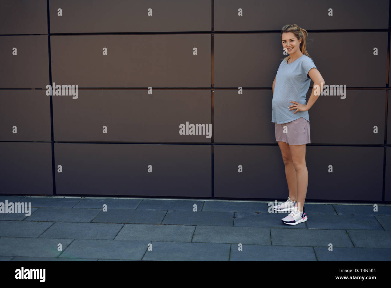 Fit athletische junge schwangere Frau in Sportkleidung vor einem Geschäftshaus mit Händen auf den Hüften in die Kamera lächelnd posiert Stockfoto
