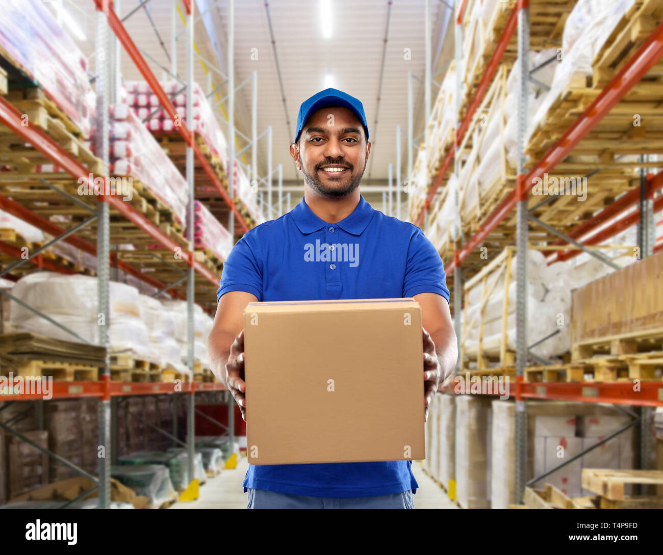 Mail Service und Versand Konzept - glückliche indischen Delivery Man oder Lagerarbeiter mit Box in der blauen Uniform über waren Hintergrund Stockfoto