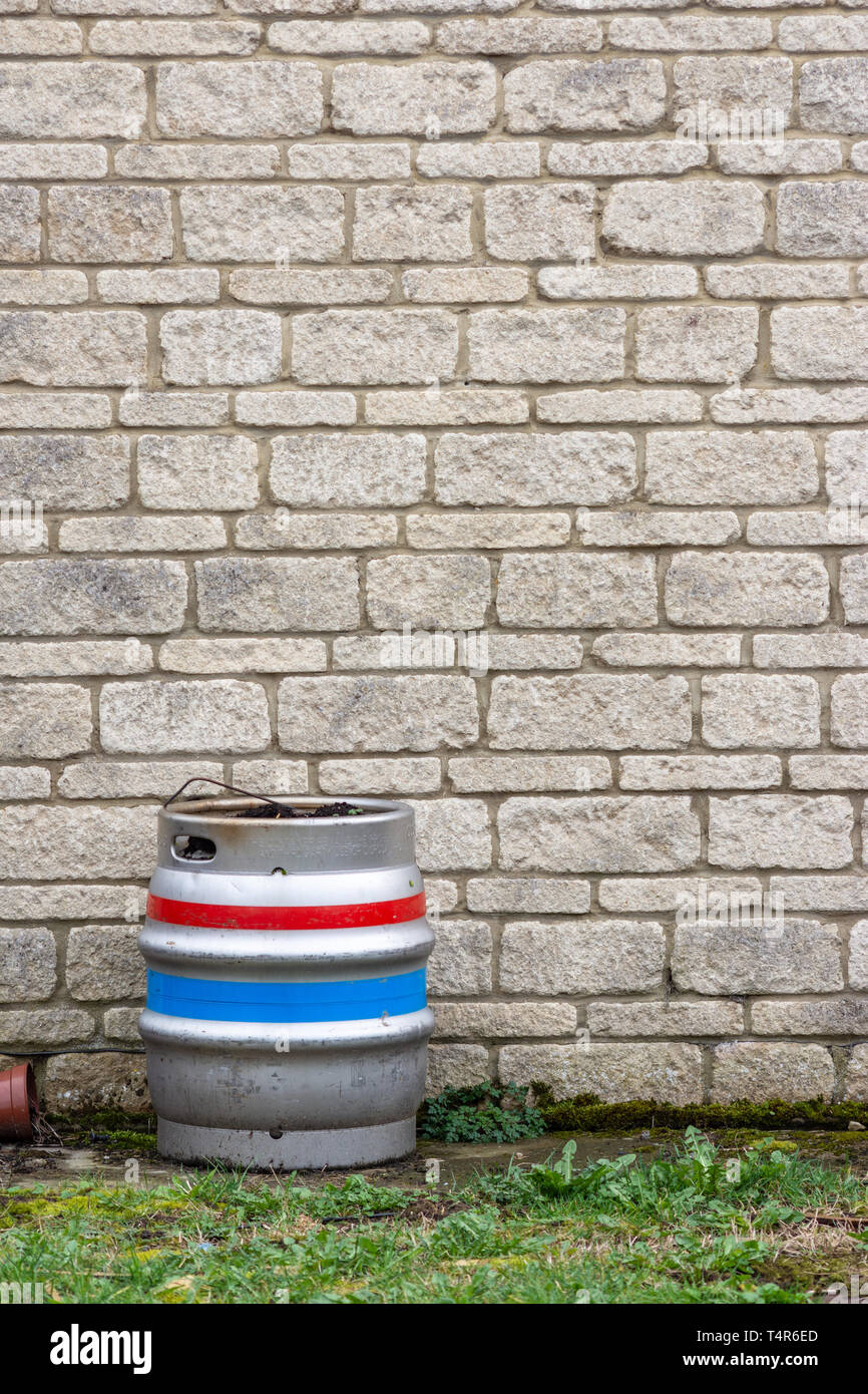 Single metal Barrel Bier Fass mit roten, weißen und blauen Streifen, stand gegen eine Wand von sandigen Stein Steine und ein Streifen von Gras im Vordergrund. Stockfoto