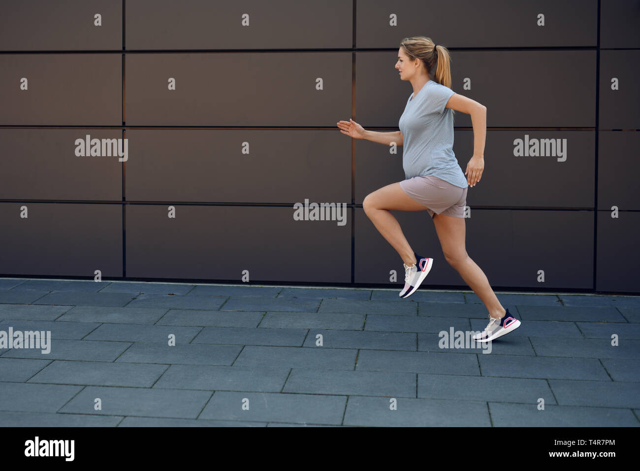 Schwangere junge Frau fit halten Sprinten entlang eines gepflasterten Gehweg vor einer Wand in einem gesunden Lebensstil Konzept Stockfoto