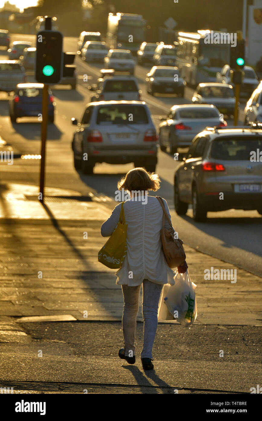 Ein Porträt einer Frau mit Einkaufstüten, Wandern entlang der Fußweg von einer geschäftigen Straße mit Hintergrundbeleuchtung bei Sonnenuntergang. Western Australia. Stockfoto