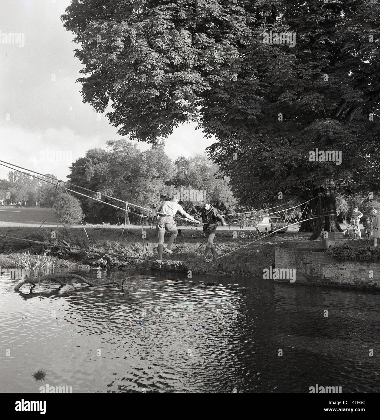 1960, historische, Scouting, team leader stehen auf einem "seilbrücke" über einen Fluß hilft einem Jungen venture Scout überqueren Sie die Brücke, England, UK. Die Venture scout Motto ist Herausforderung. von Robert Baden-Powell Gegründet 1907, die Pfadfinderbewegung hat einen starken Fokus auf die Natur und das Überleben Fähigkeiten, mit dem Ziel jungen Menschen physischen, psychischen und geistigen Entwicklung zu unterstützen. Stockfoto