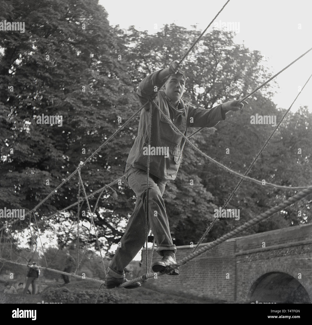 1960, historische, eine Venture Scout einen Fluß überquert auf einer Seilbrücke, England, UK. Das partnerunternehmen unter dem Motto "Challenge". Von Robert Baden-Powell Gegründet 1907, die Pfadfinderbewegung hat einen starken Fokus auf die Natur und das Überleben Fähigkeiten, mit dem Ziel jungen Menschen physischen, psychischen und geistigen Entwicklung zu unterstützen. Stockfoto