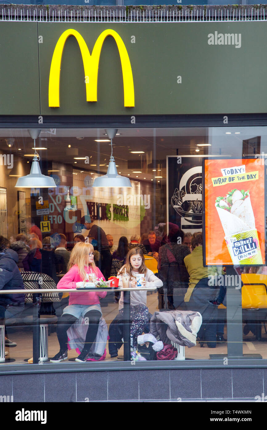 Zwei junge Mädchen sitzen, Essen und Trinken im Fenster des Fast Food von McDonald's in der High Street Shop Stockfoto