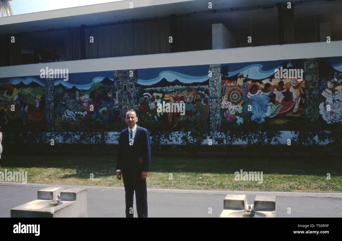 Ein Mann steht in drei-viertel länge, vor einem Wandbild posiert, wahrscheinlich auf dem Zentralamerika und Panama Pavillon, an einem sonnigen Tag, in New York World's Fair, Flushing Meadows Park, Queens, New York, Juni 1964. () Stockfoto