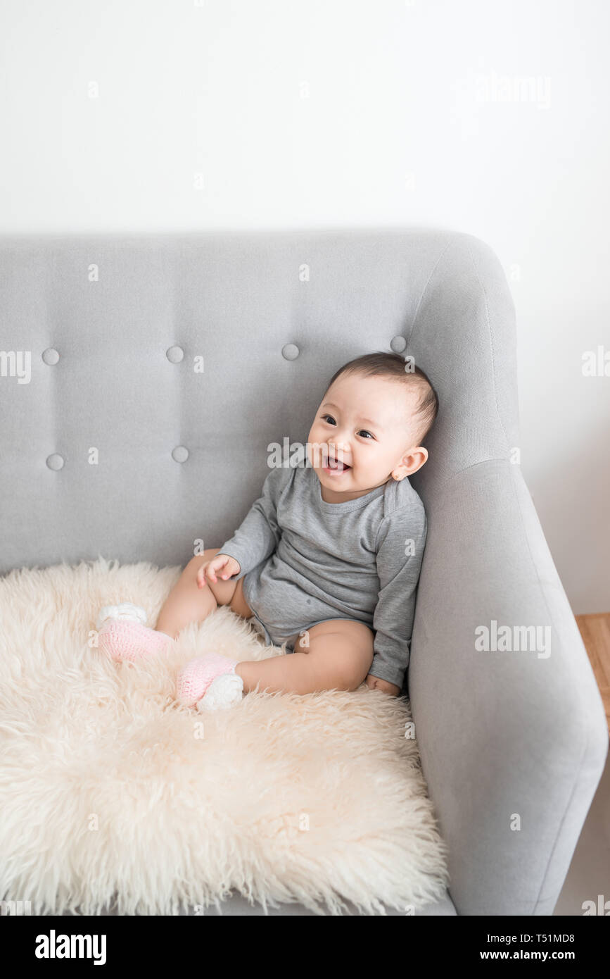 Closeup Portrait von cute adorable Lächeln Lachen, Baby Mädchen mit schwarzen Augen sitzen auf einem Sofa weg von der Kamera suchen, natürliche Fenster Licht, lifestyl Stockfoto