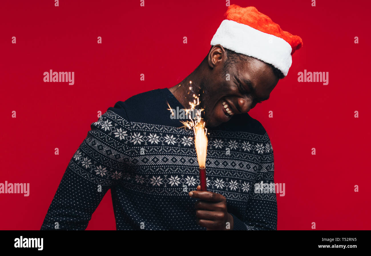 Fröhlicher junger afrikanischer Mann mit Wunderkerzen auf rotem Hintergrund. Jungen Kerl trägt einen santa hut mit Feuerwerk. Stockfoto