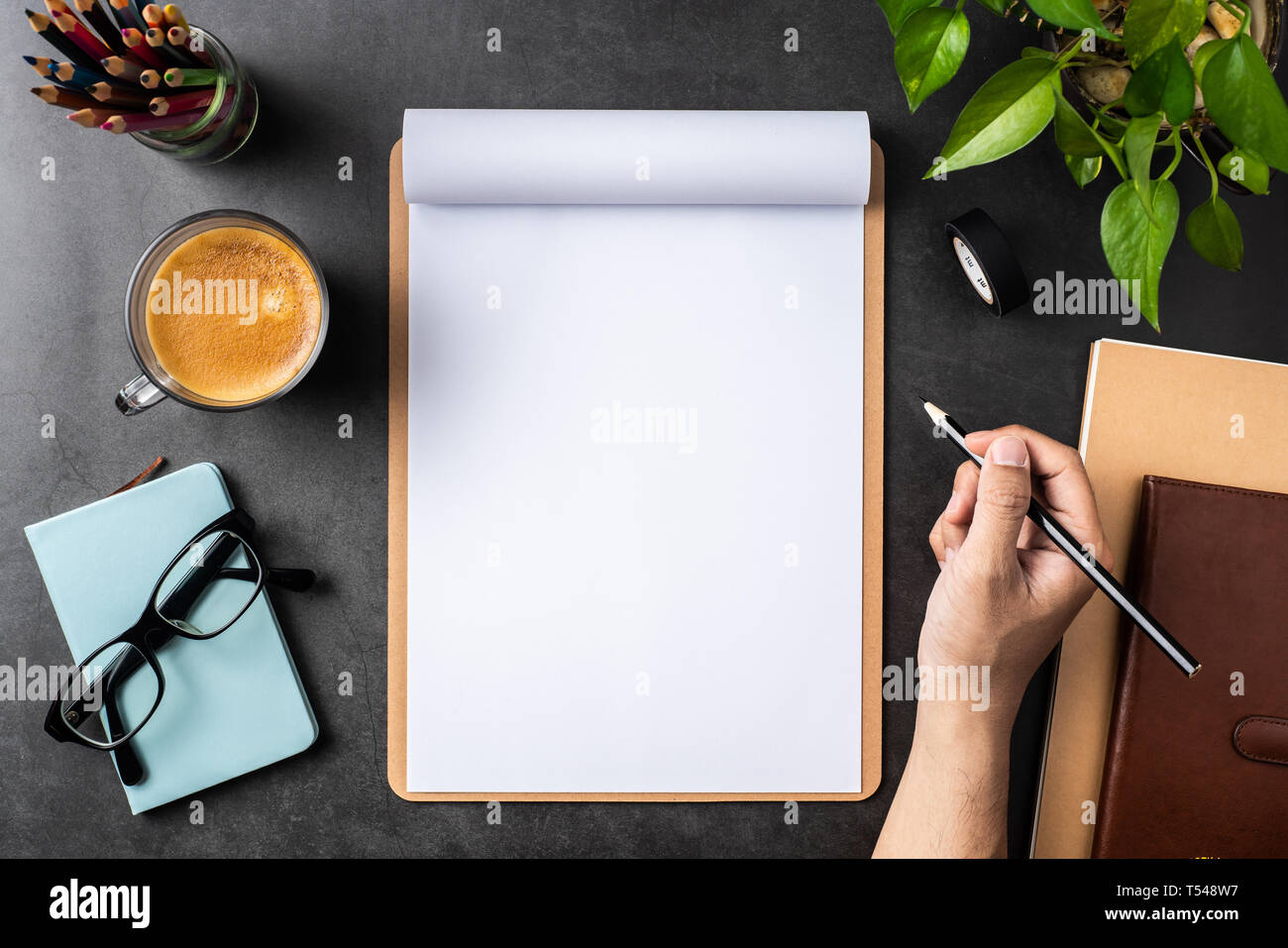 Designkonzept - Blick von oben auf die Hand halten Bleistift auf weißem A4-Papier umgedreht, mit braunen Zwischenablage, Gläser, Topfpflanze, Kaffee, Bleistift auf Schwarz hinterg Stockfoto