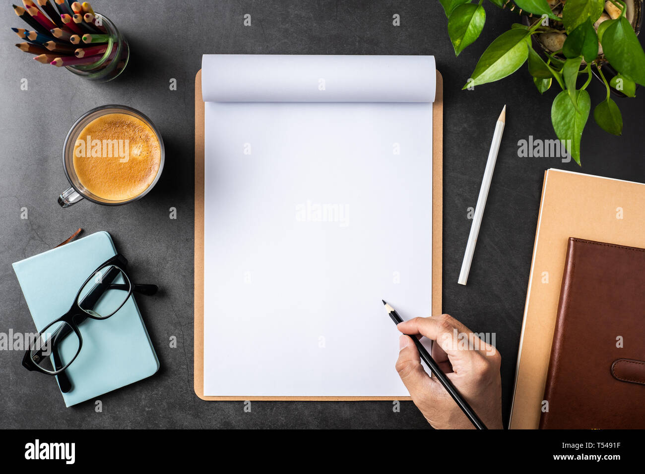 Designkonzept - Blick von oben auf die Hand halten Bleistift auf weißem A4-Papier umgedreht, mit braunen Zwischenablage, Gläser, Topfpflanze, Kaffee, Bleistift auf Schwarz hinterg Stockfoto