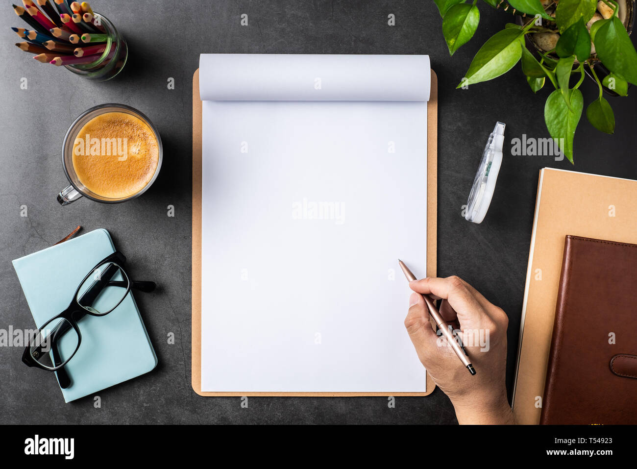 Designkonzept - Blick von oben auf die Hand halten Bleistift auf weißem A4-Papier umgedreht, mit braunen Zwischenablage, Gläser, Topfpflanze, Kaffee, Bleistift auf Schwarz hinterg Stockfoto