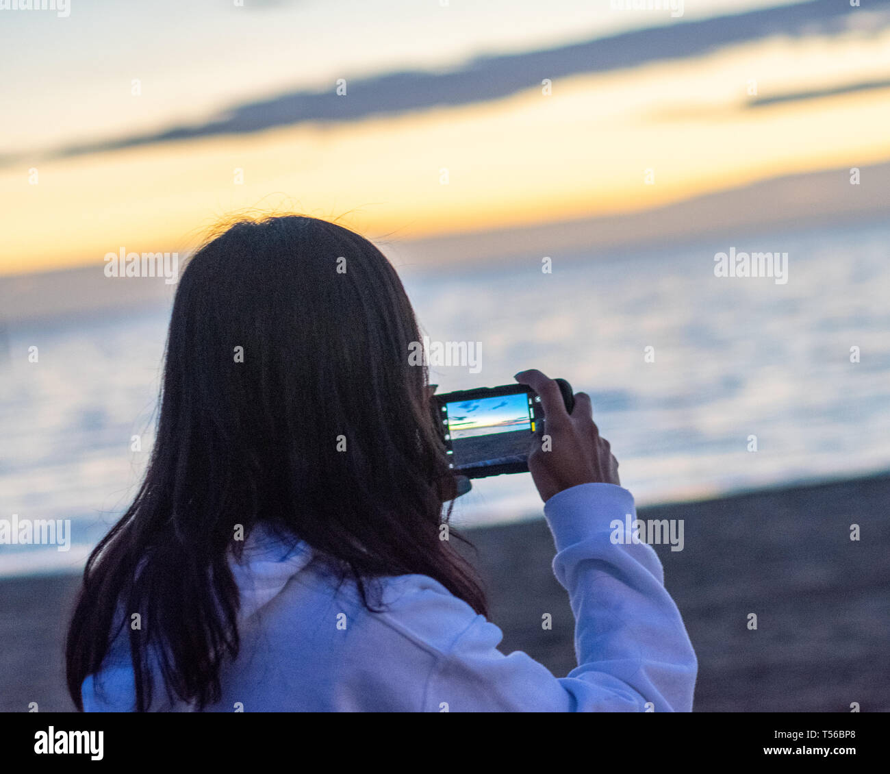 Mädchen ein Bild von den Sonnenuntergang am Strand mit Ihrem Mobiltelefon von hinten Stockfoto