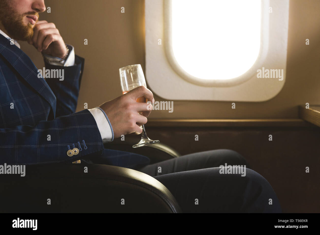 Ein junger erfolgreicher Geschäftsmann in einer teuren Anzug sitzt in den Stuhl eines privaten Jet mit einem Glas Champagner in der Hand und schaut aus dem Fenster Stockfoto