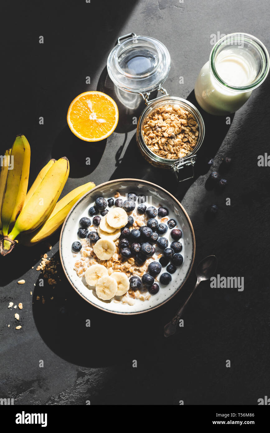 Gesundes Frühstück Müsli Schale mit Banane, Blaubeere und Milch auf einem schwarzen konkreten Hintergrund. Ansicht von oben. Grelles Licht Foodfotografie Stockfoto