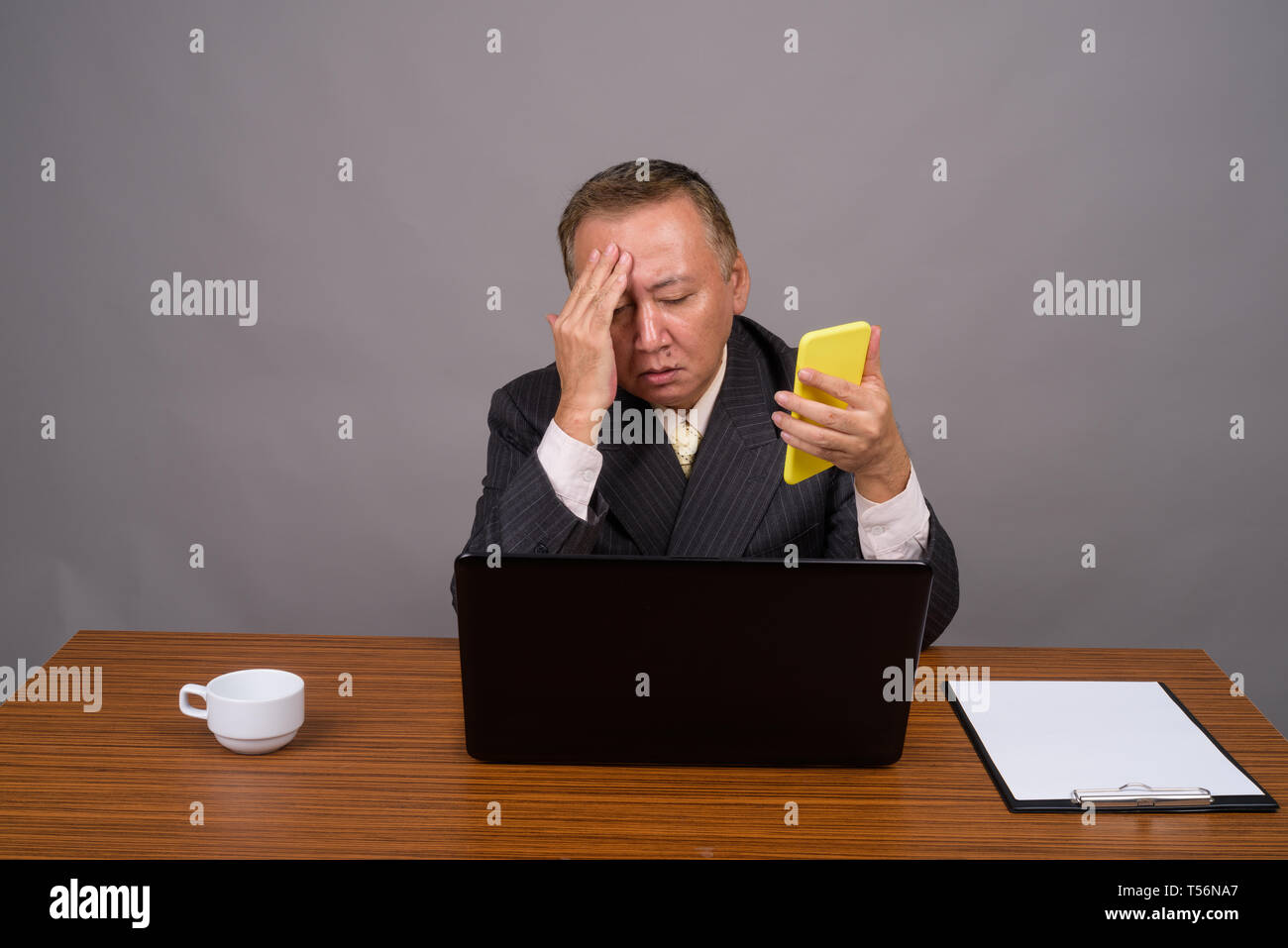 Reifen asiatische Geschäftsmann sitzen mit hölzernen Tisch gegen Grau Stockfoto