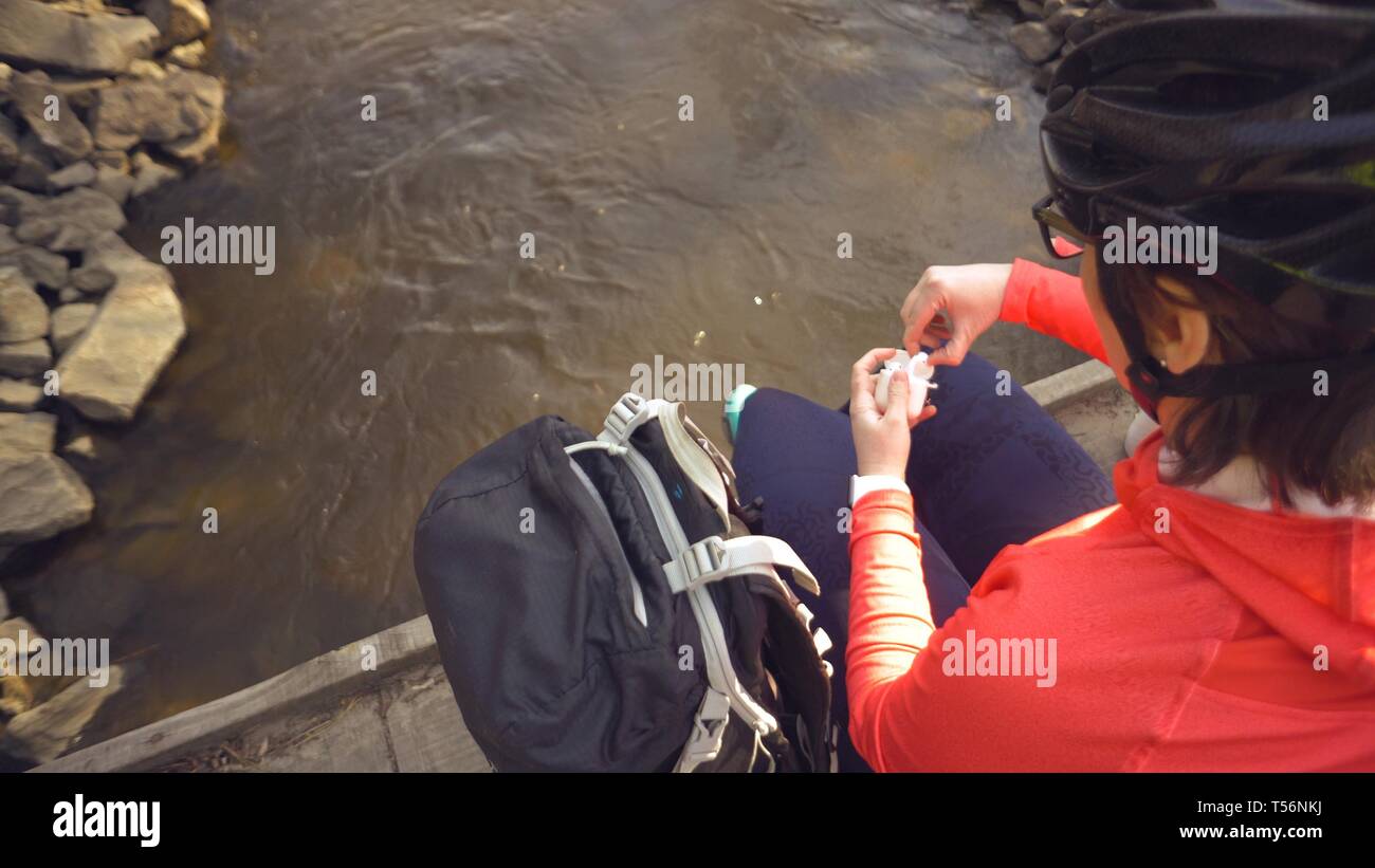 Kaukasische Frau Sportler in einem hellen Sportswear Helm ruht auf einer Brücke über den Fluss. Radfahrer mit weißen Taschen, das Gehäuse zu öffnen, legen Sie Stockfoto