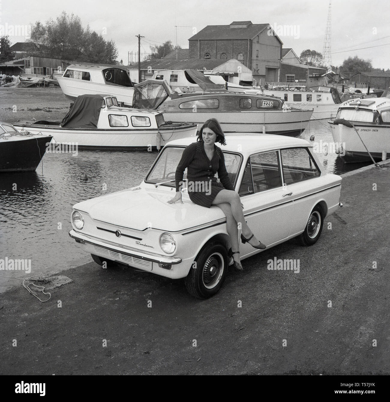 1967, historisch, draußen in einer Werft, sitzt eine junge Dame mit Wolljacke und Rock auf dem Kofferraum eines Sunbeam Sport Motorwagens, der Sportversion des berühmten Kleinwagens Hillman Imp. Der Hillman Imp war ein Kleinwagen der Rootes Group und deren Nachfolger Chrysler Europe von 1963 bis 1976. Er war der Konkurrent in der Kleinwagenklasse des britischen Leyland Mini. Stockfoto