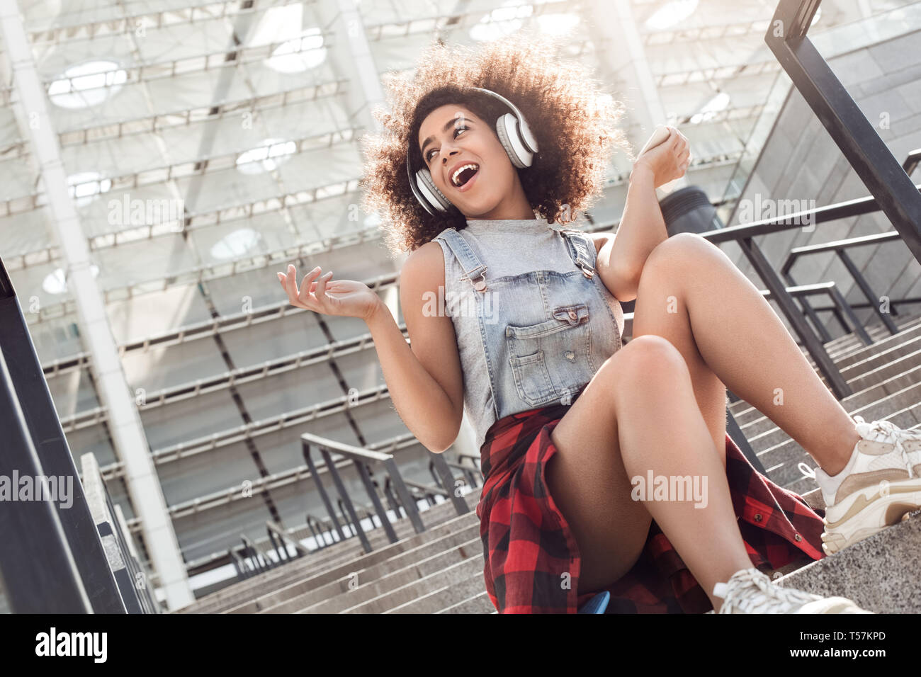 Junge stilvolle Frau in der Stadt Straße sitzen auf betontreppen mit Kopfhörern Musik hören Holding smartphone singen fröhlich Suchen asid Stockfoto